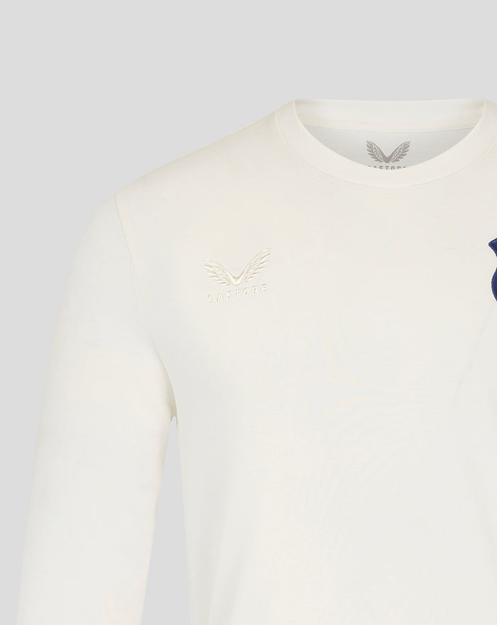 Close up of cream long sleeve shirt showing embossed Castore logo on left chest and blue Wembley crest on right chest.
